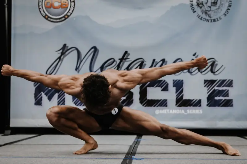 Austin Green on stage in black posing trunks, low in a wide spread pose with arms stretched out.